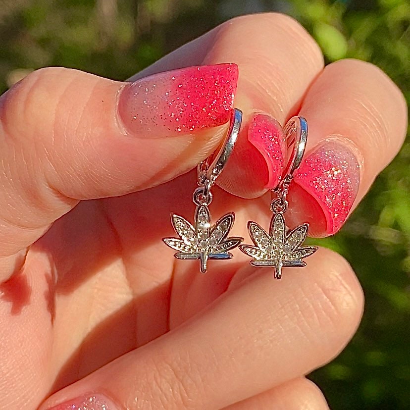 two marijuana symbol drop earrings with cubic zirconia detailing and silver hardware held in hand